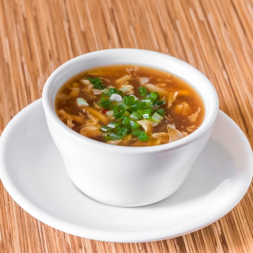 A white bowl of hot and sour soup, garnished with chopped green onions, on a wooden table.